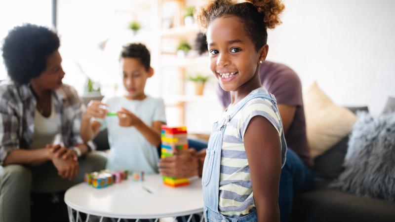 Smiling young girl standing in front of table with family playing game behind her