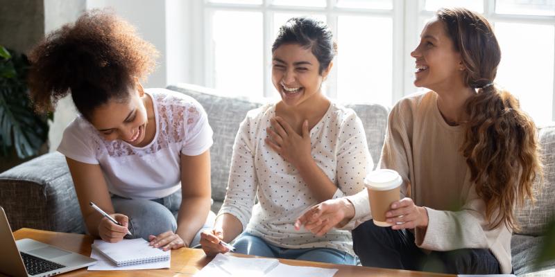 Three diverse teen females sitting on couch laughing and doing homework